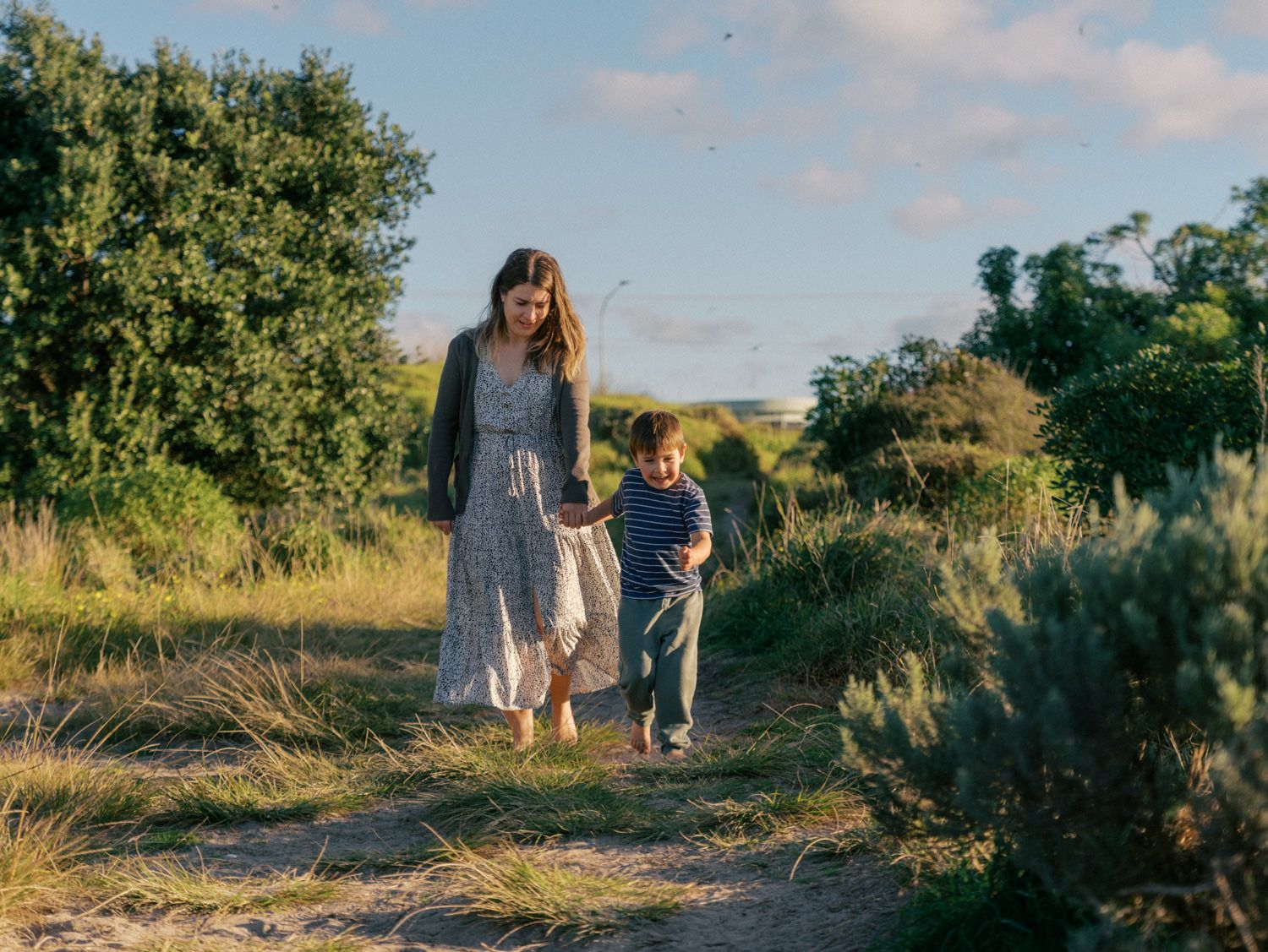 A mother and young child walk hand in hand along a dirt path surrounded by lush green trees and grass.