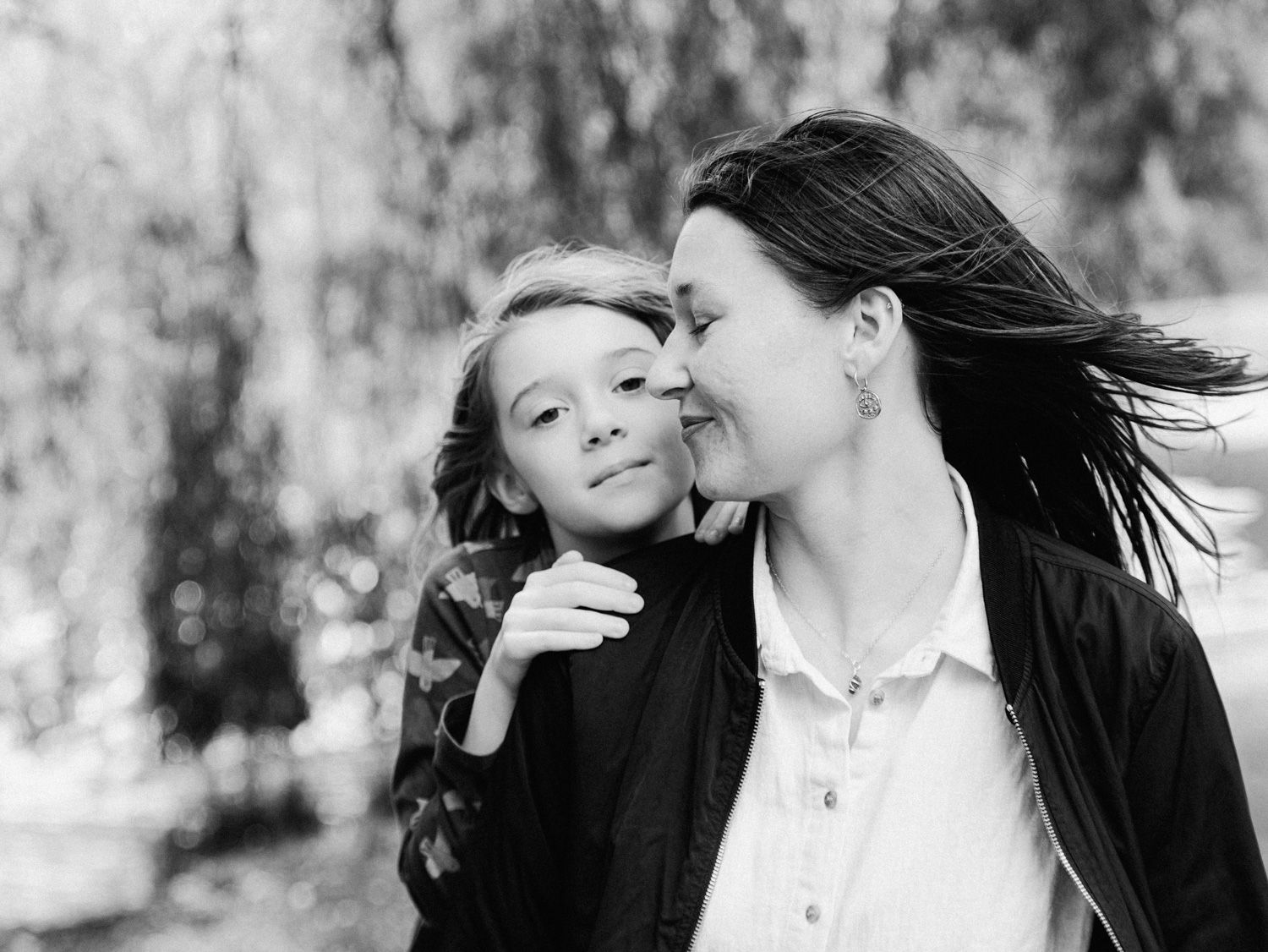 Black and white photo of a mother tenderly kissing her young daughter outdoors near willow trees.