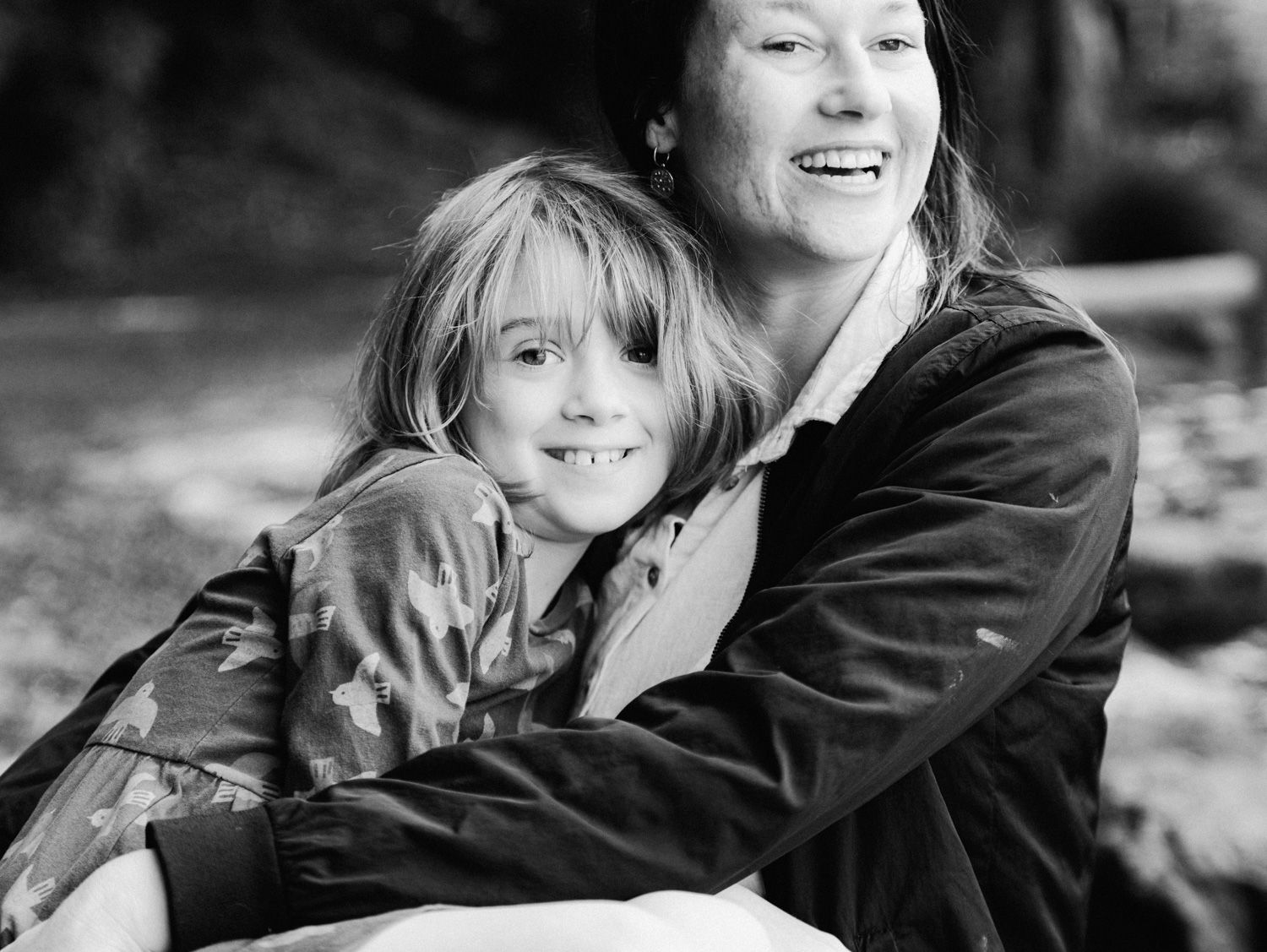Black and white photo of a smiling mother hugging her young blonde daughter outdoors.