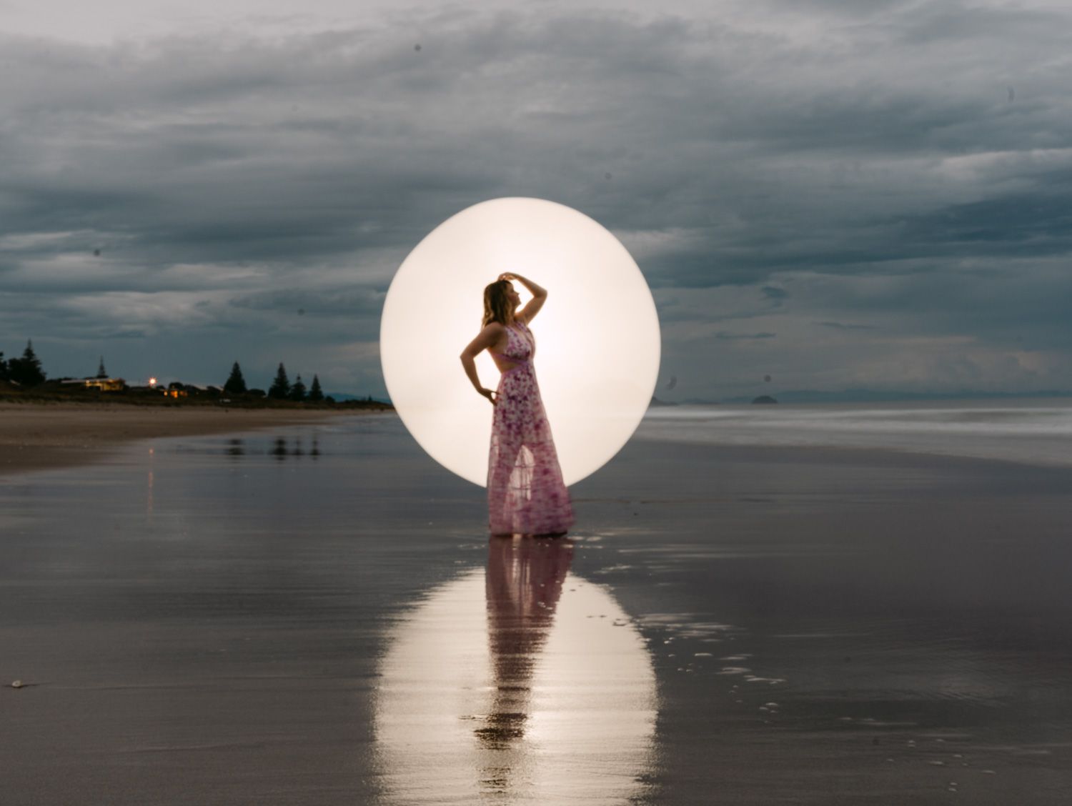 Woman in floral dress standing on reflective beach before glowing circle, dramatic cloudy sky backdrop.