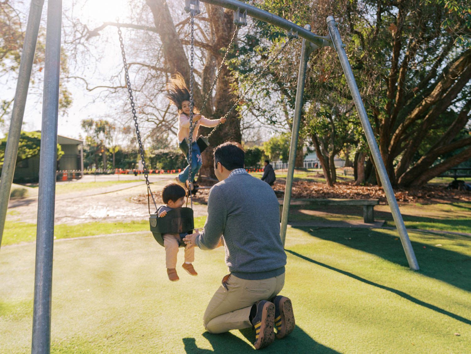 Father plays with two children on a swing set at a sunny park playground on green grass.
