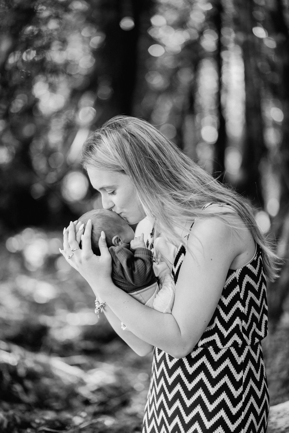 Black and white photo of a mother tenderly kissing her newborn baby outdoors in a forest setting.