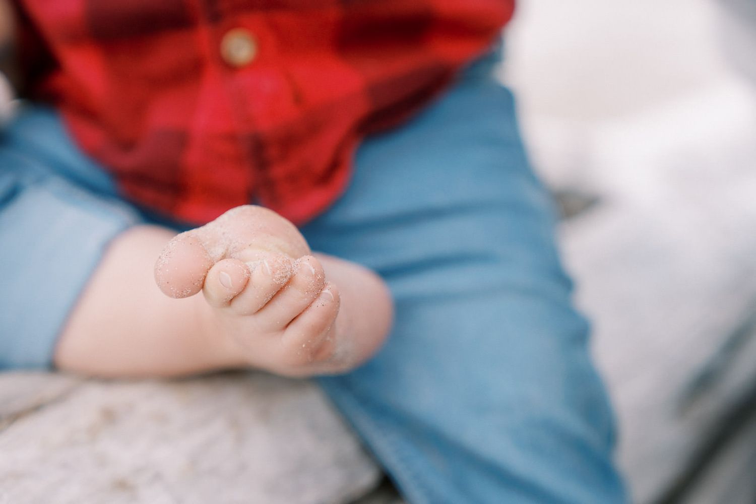 Close-up of a baby's tiny bare foot and toes, wearing red plaid shirt and blue jeans, sitting on a soft surface.