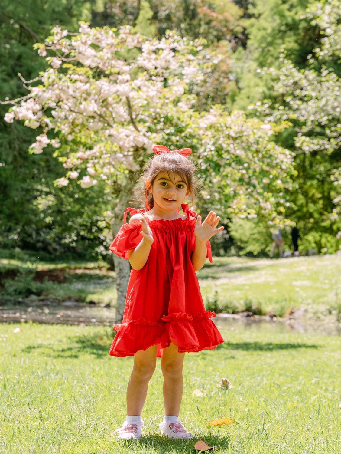 A young girl in a red dress and bow stands on grass near a flowering tree in a sunny park.