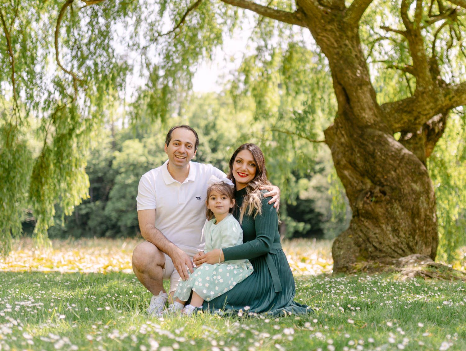 Happy family of three posing outdoors under a willow tree in a flower-filled meadow on a sunny day.