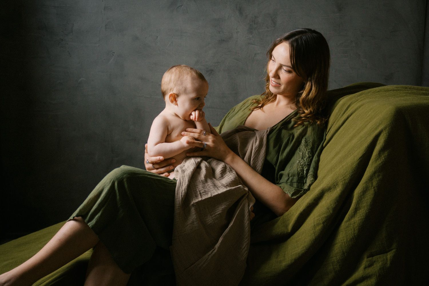 Mother in green dress sits on sofa holding baby wrapped in olive blanket in moody studio setting.