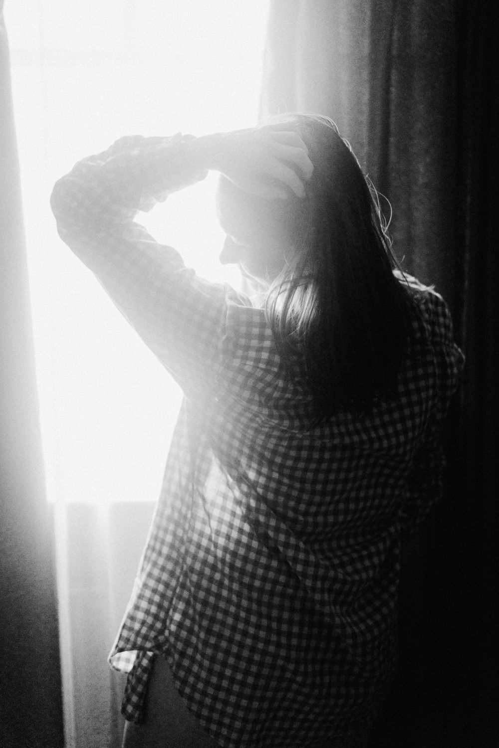 Black and white photo of a woman in a plaid jacket standing by a bright window with her hand raised to her head.