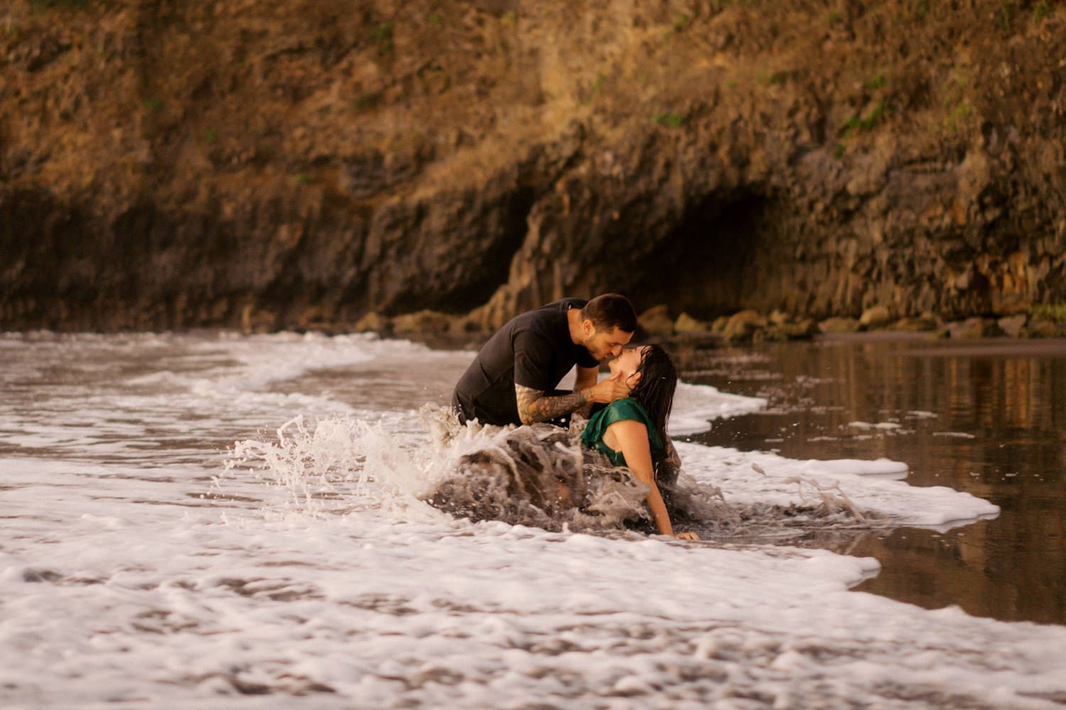 Couple sharing a romantic kiss in ocean waves on a rocky beach, wearing black and green outfits.
