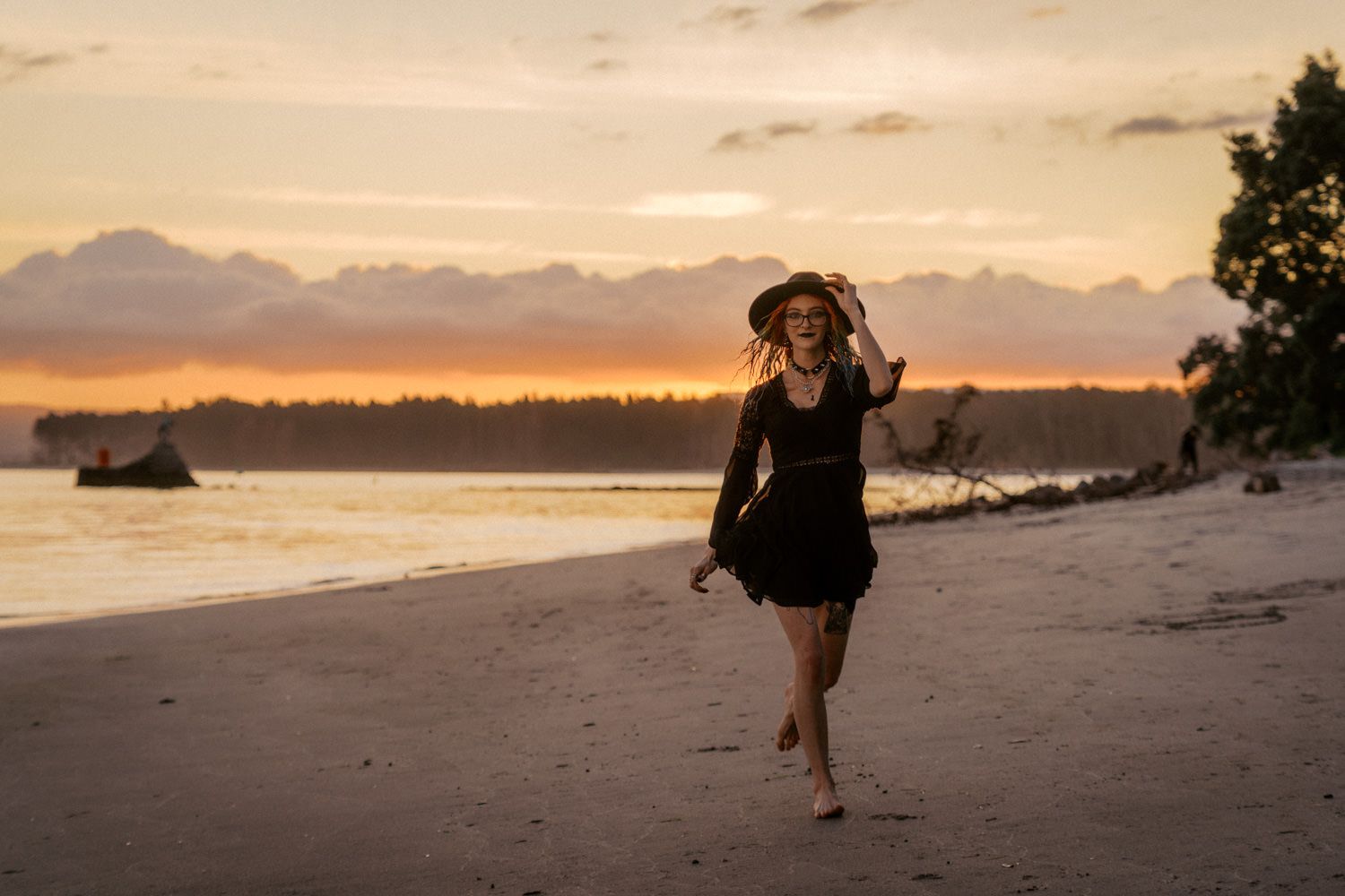 Woman in black dress and hat walks barefoot on sandy beach during golden sunset with dramatic clouds.