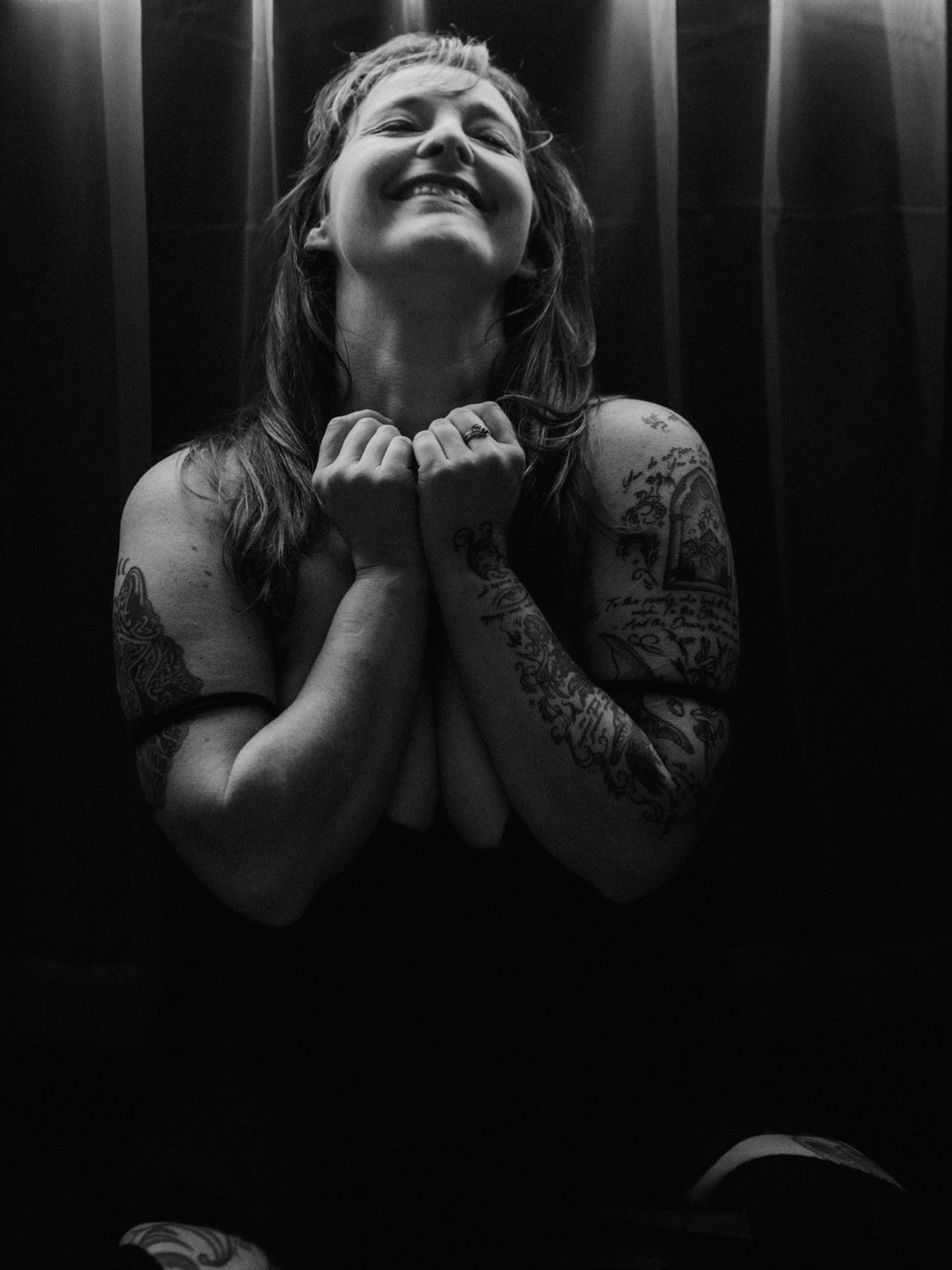 Black and white photo of tattooed woman smiling with hands clasped in prayer pose against dark striped background.