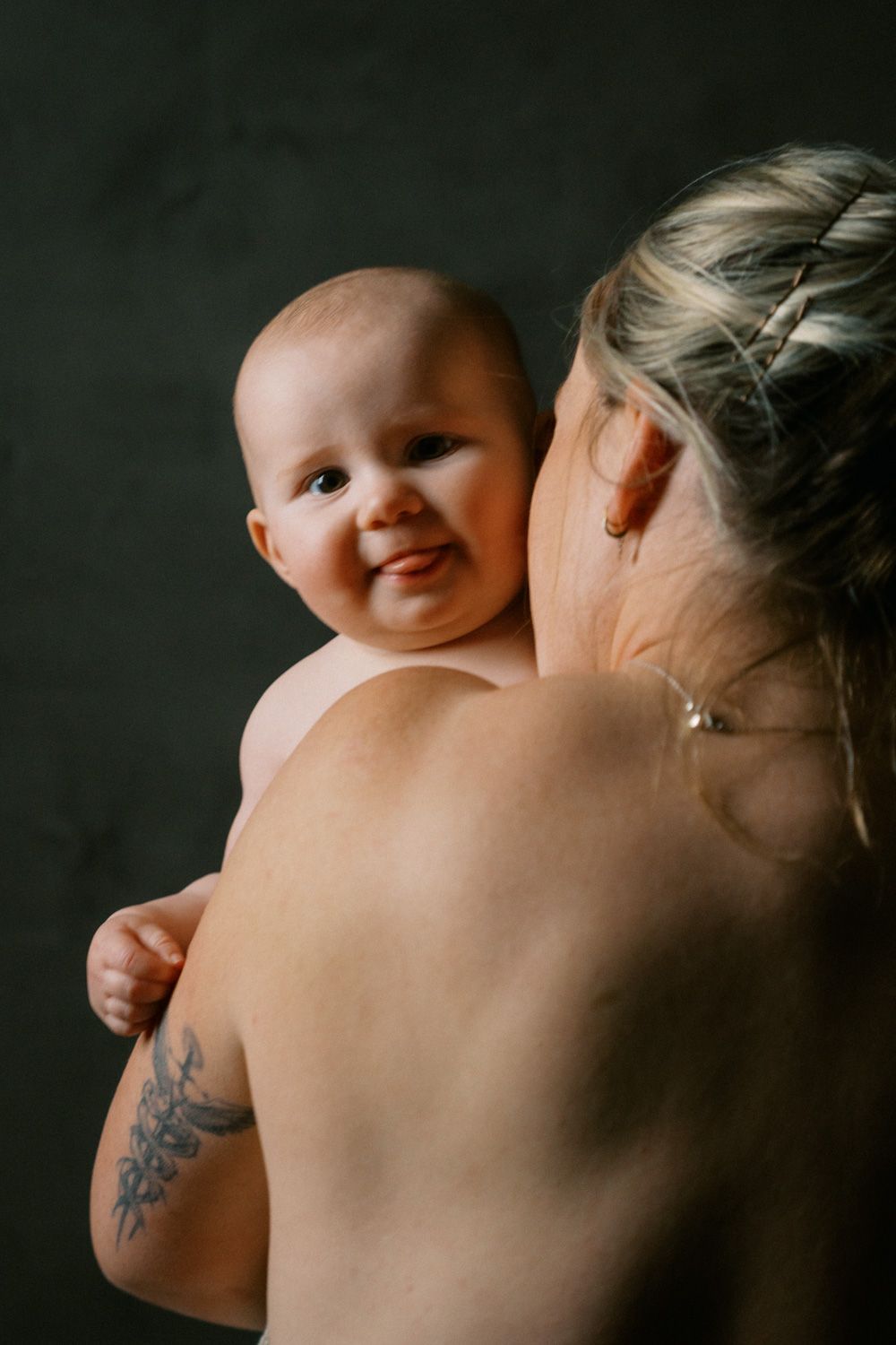 A smiling baby being held over the shoulder of a tattooed woman with braided blonde hair against a dark background.