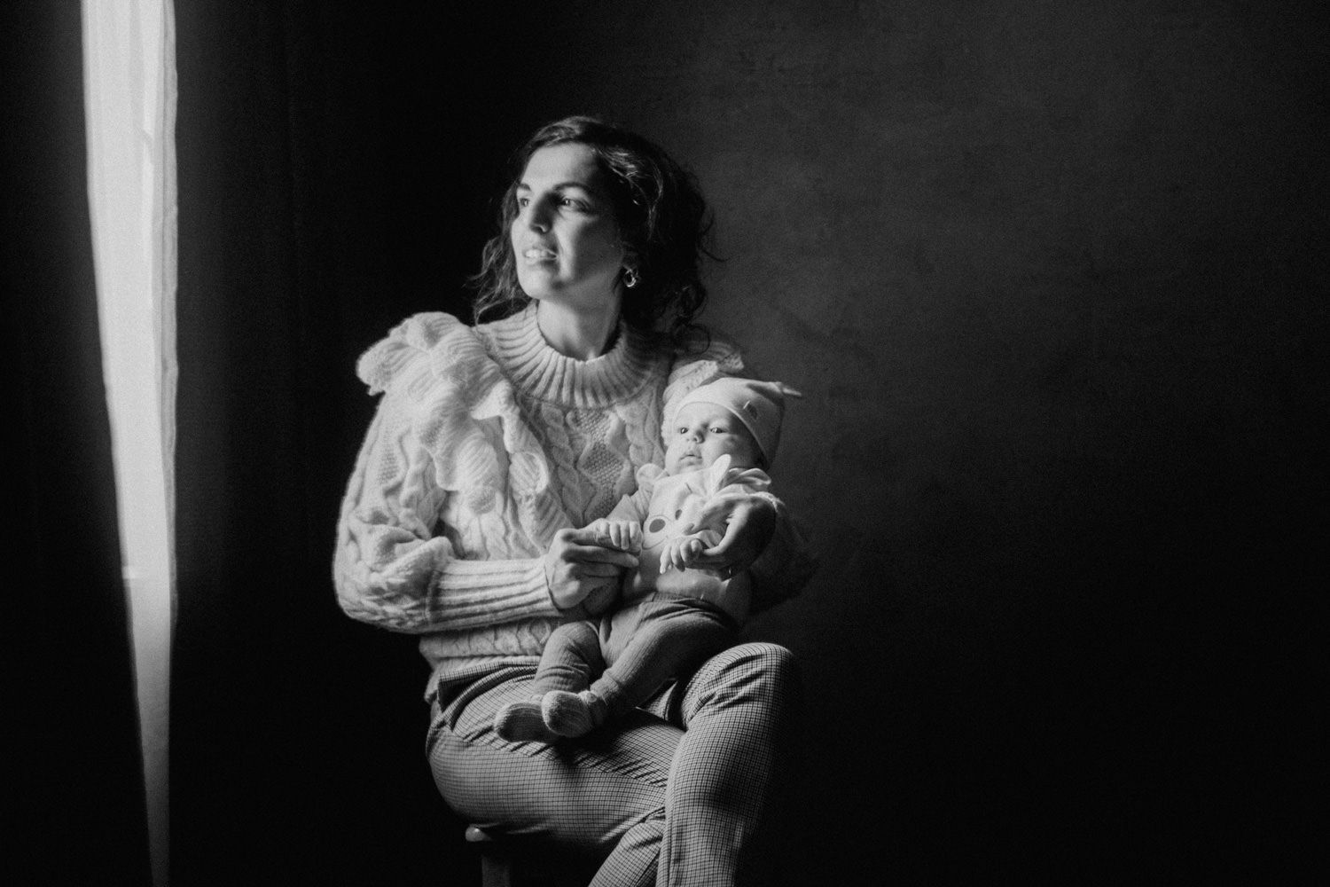 Black and white portrait of a woman sitting and cradling a baby, lit by soft window light.