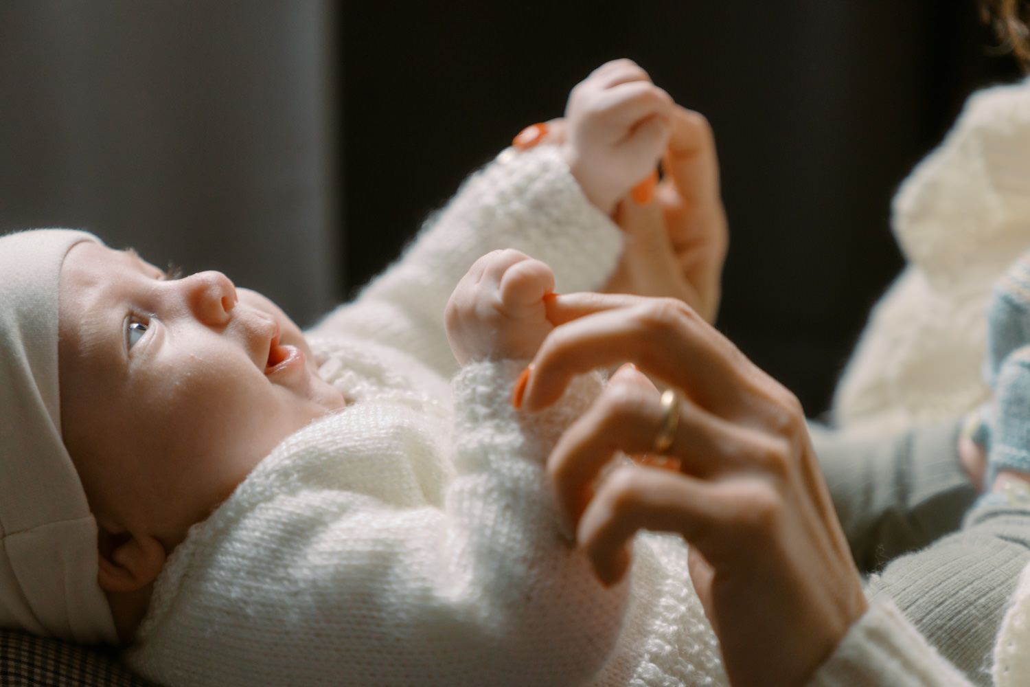 A newborn baby in a white knit outfit and hat lies on their back while an adult gently holds their tiny hands.