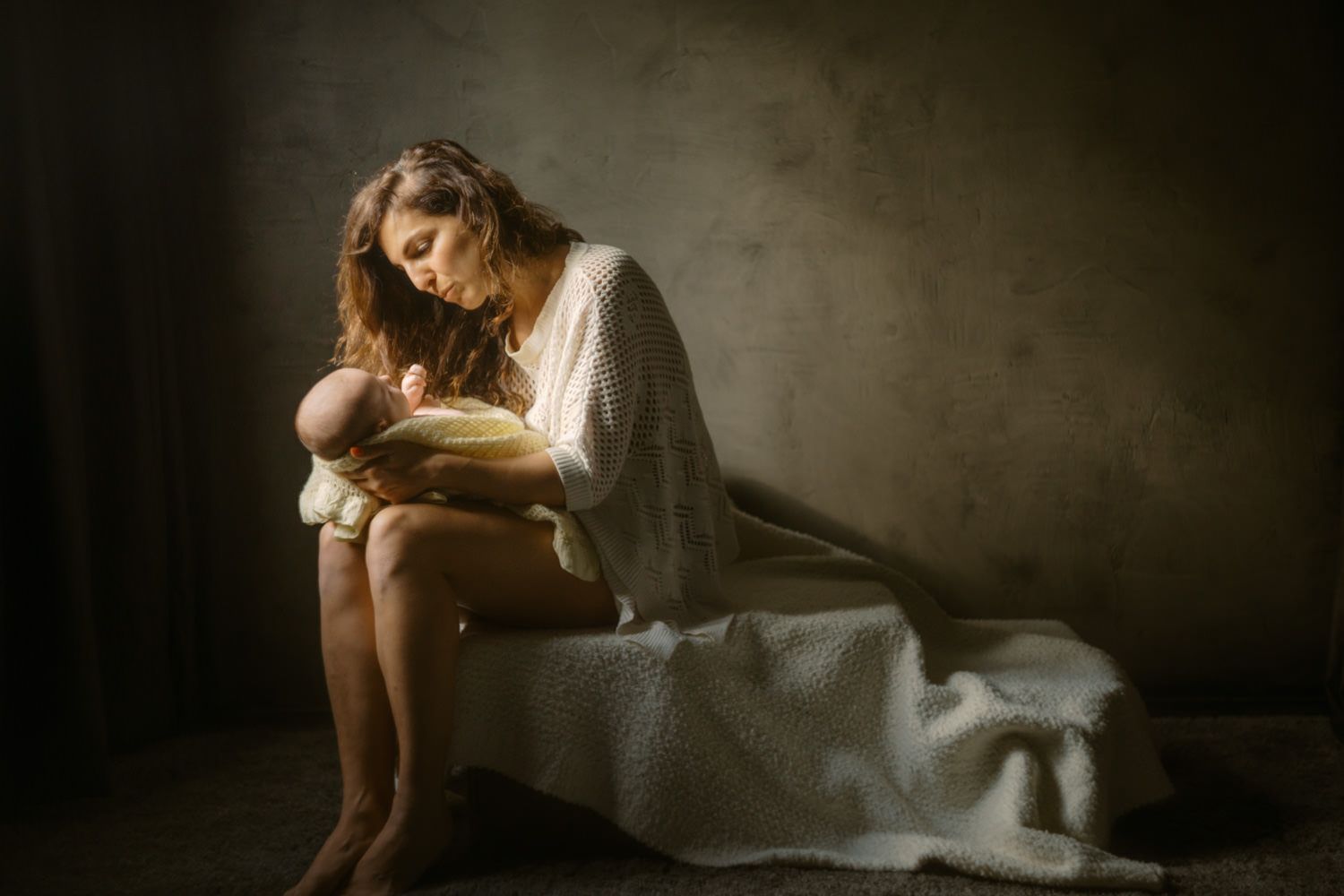 A young mother in a white dress tenderly holds her newborn baby while seated in soft, dramatic lighting.