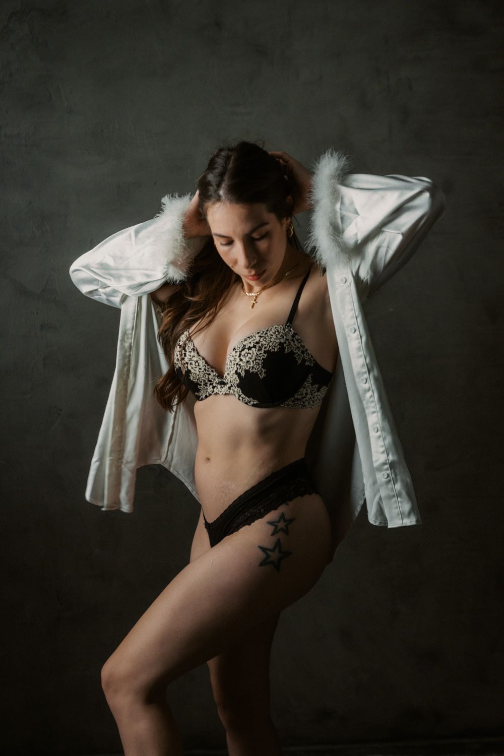 Woman in black lace lingerie set with open white feathered shirt, star tattoo on thigh, dark background.