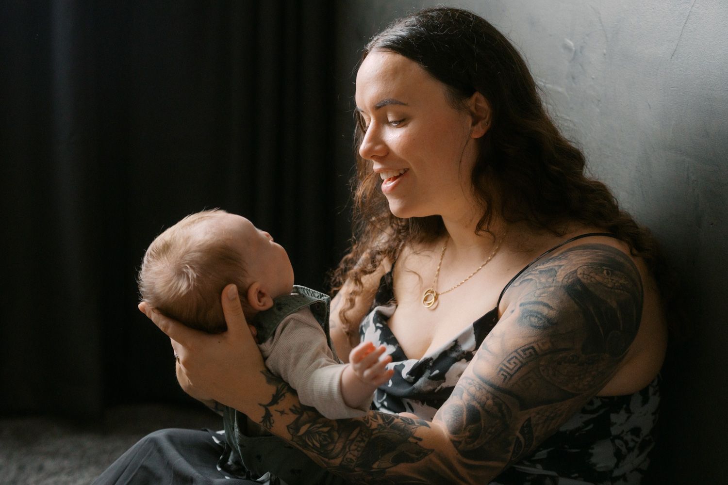 Tattooed mother smiling while holding her newborn baby against a dark background.