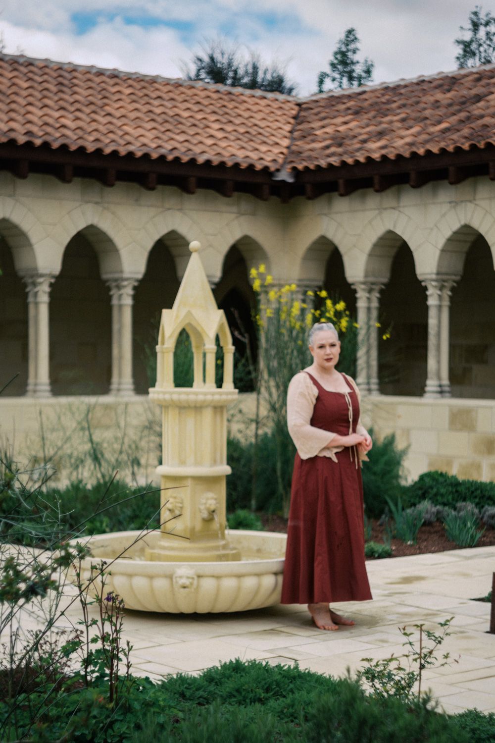 Woman in medieval dress stands by Gothic fountain in arched stone courtyard with terracotta roof.