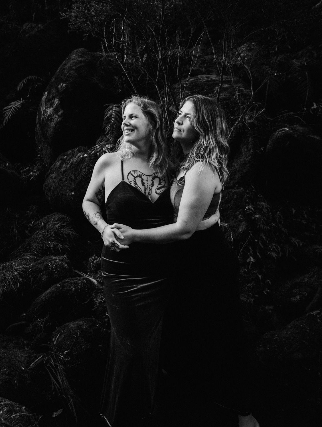 Two women in black dresses embrace and laugh together against a lush dark forest backdrop in a black and white photo.