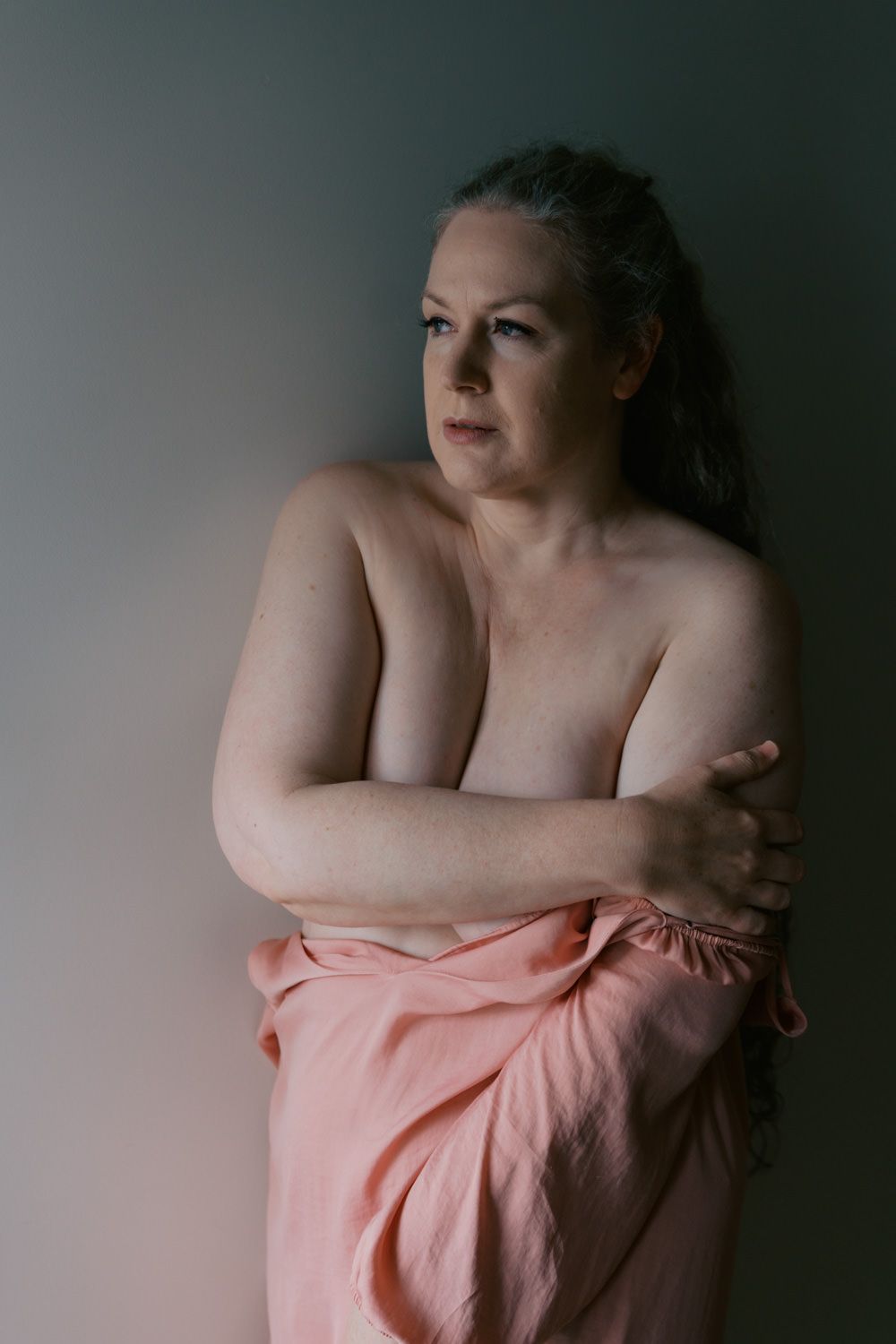 A mature woman with gray-streaked hair poses thoughtfully, draped in a pink fabric against a muted green background.