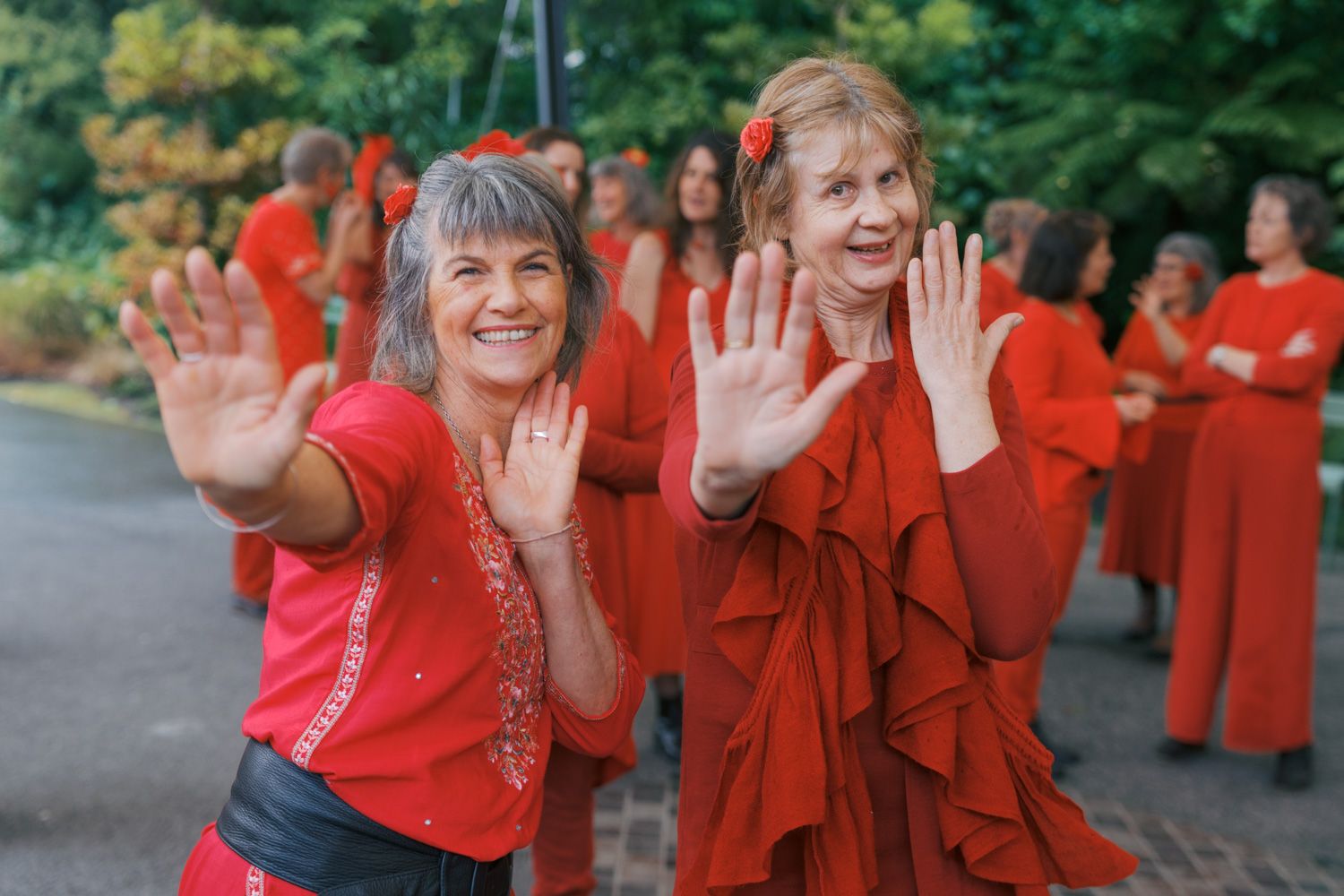 Two smiling women in red outfits wave at camera, surrounded by group dressed in red outdoors.