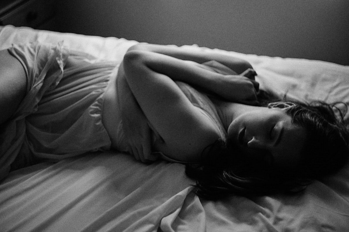 Black and white photo of a woman lying on a bed wrapped in white sheets in a moody, artistic pose.