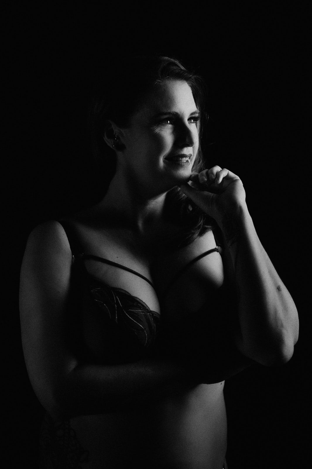 Black and white boudoir portrait of a woman in lingerie posing thoughtfully against a dark background.