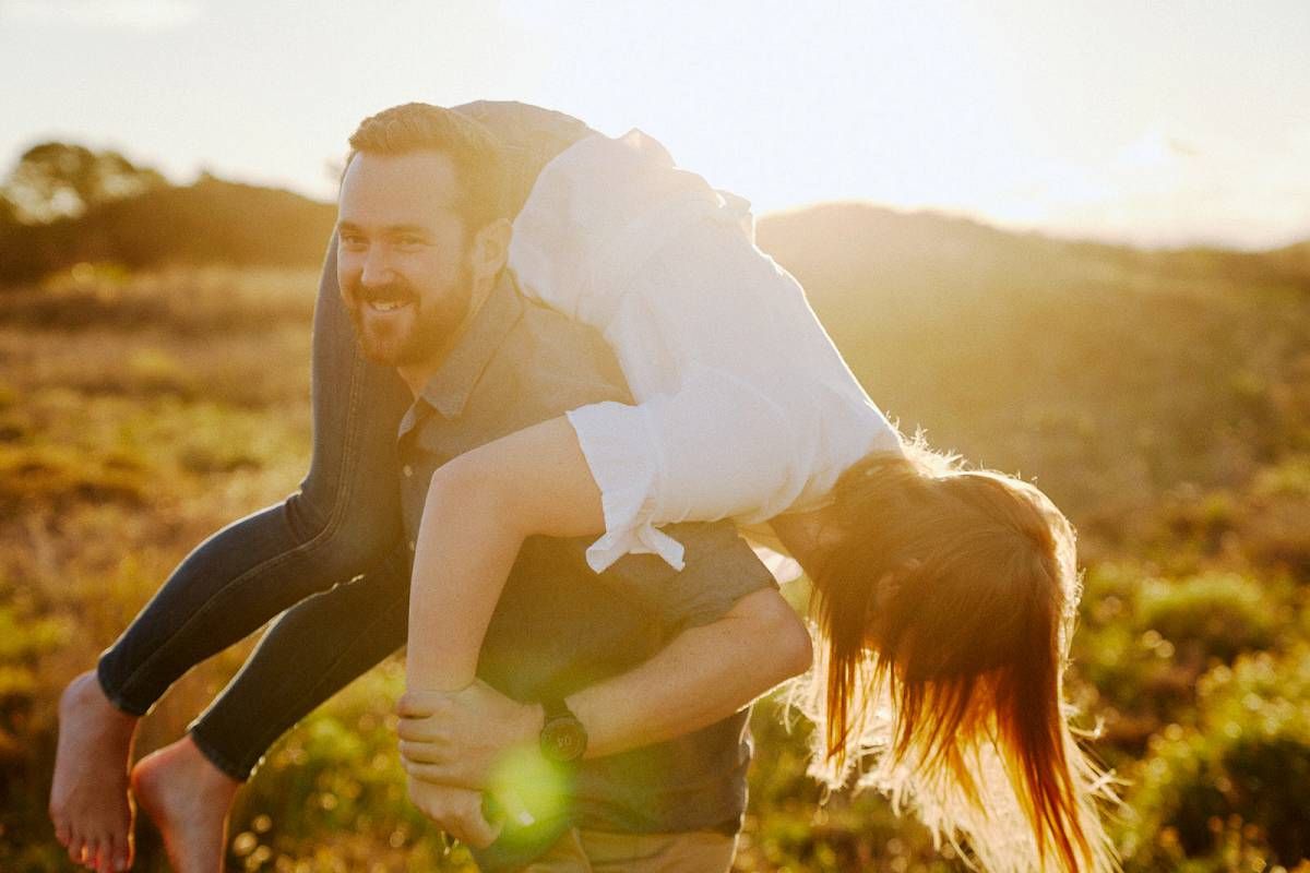 A joyful couple plays outdoors at golden hour, man lifting woman in a sunlit field with hills in the background.