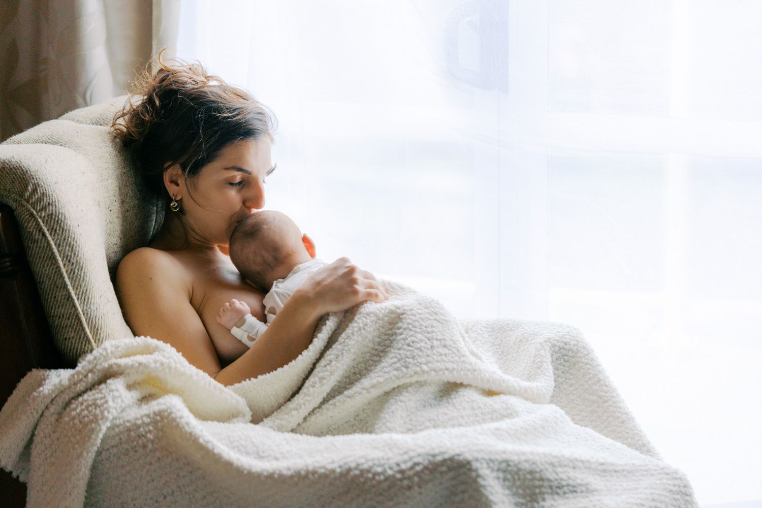 A mother tenderly holds her newborn baby wrapped in a white blanket while sitting in a chair by a bright window.