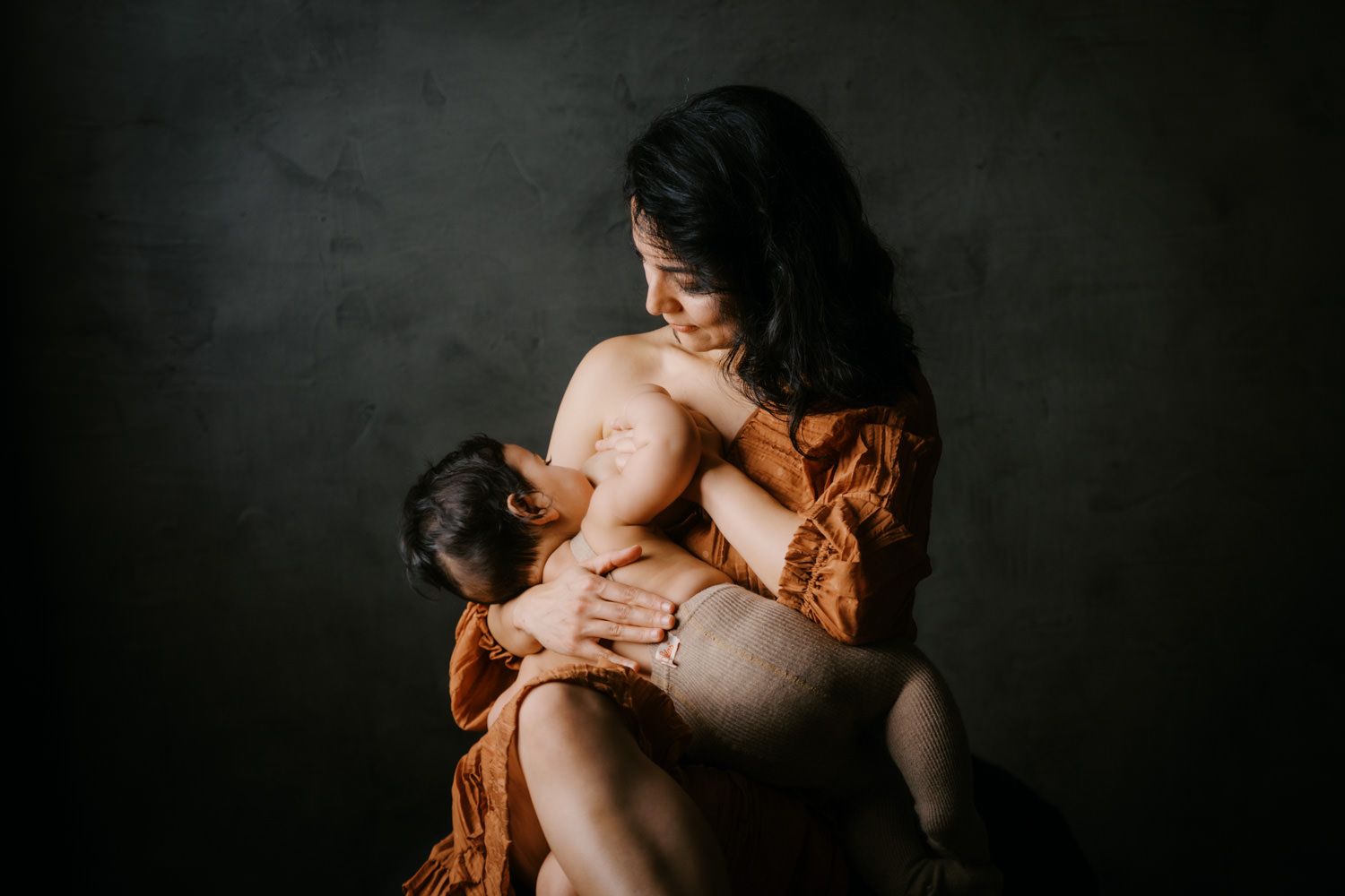 A mother in an orange wrap tenderly breastfeeds her baby against a dark background.