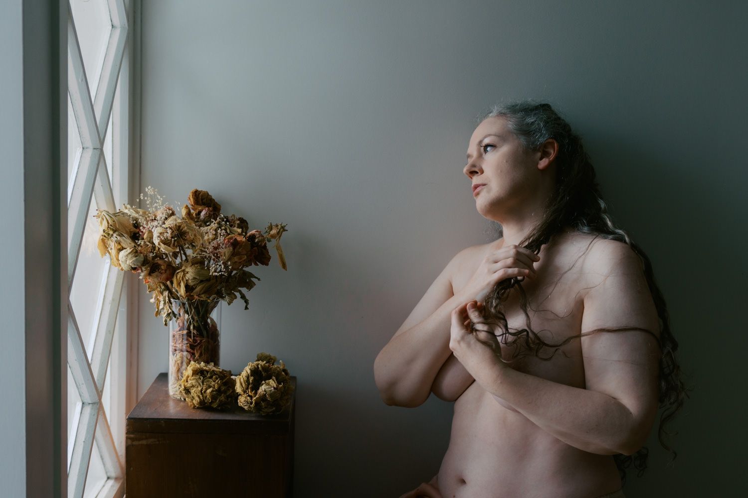 A woman with blue hair stands by a window holding dried flowers, with a floral arrangement on a wooden table nearby.