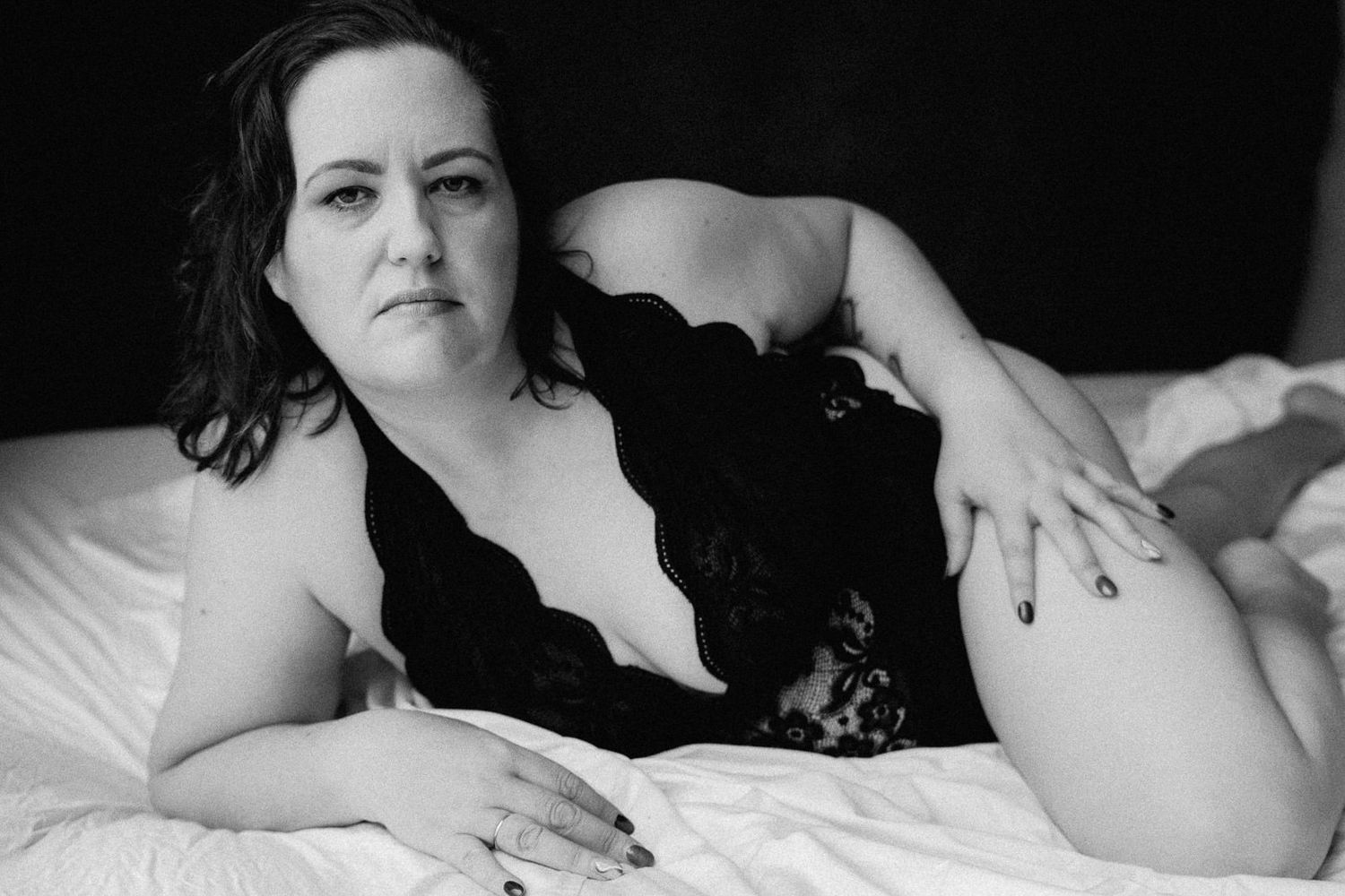 Black and white boudoir photo of a woman in lace lingerie reclining on white bedding.