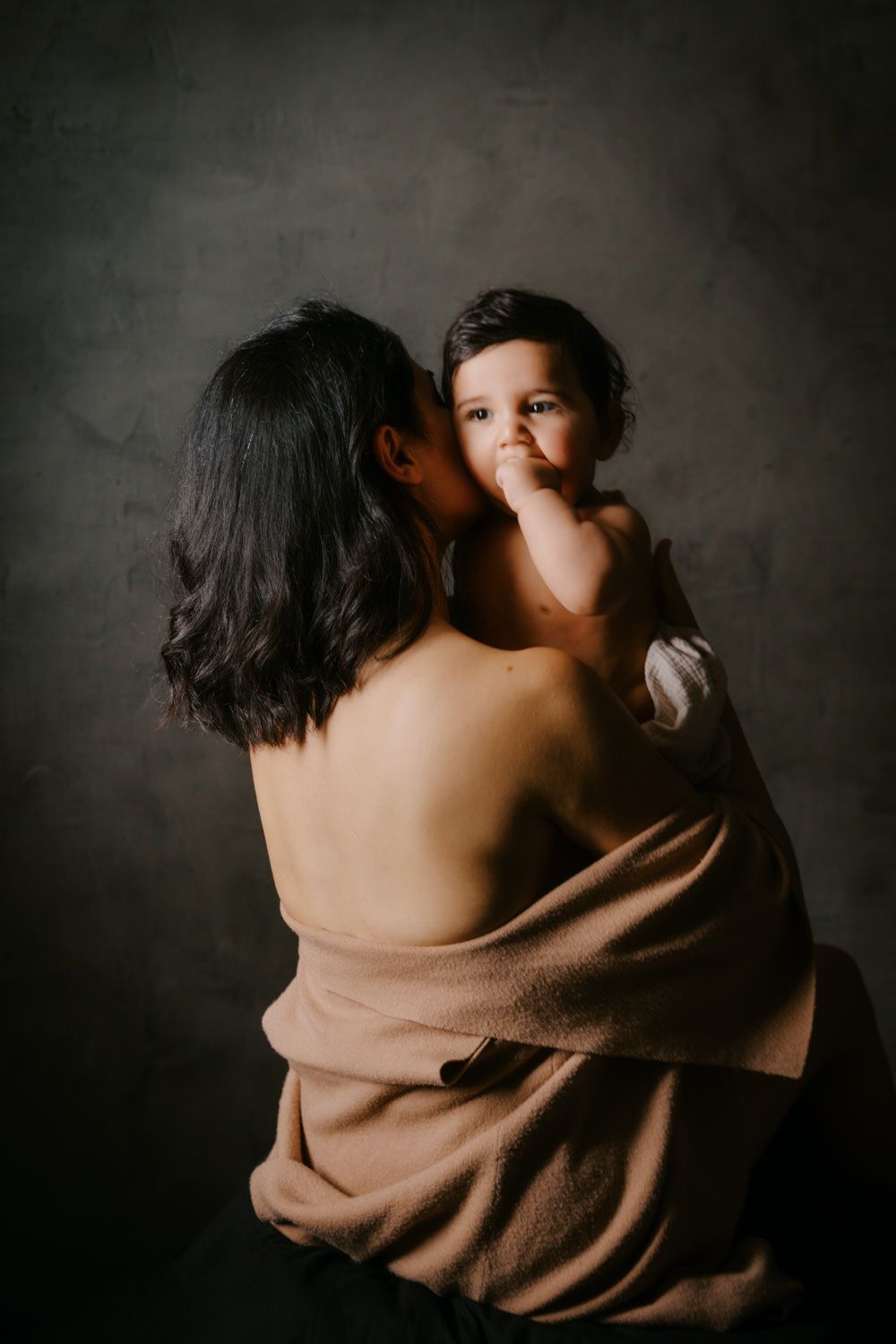 A mother wrapped in a brown towel tenderly kisses her baby in a moody studio portrait.