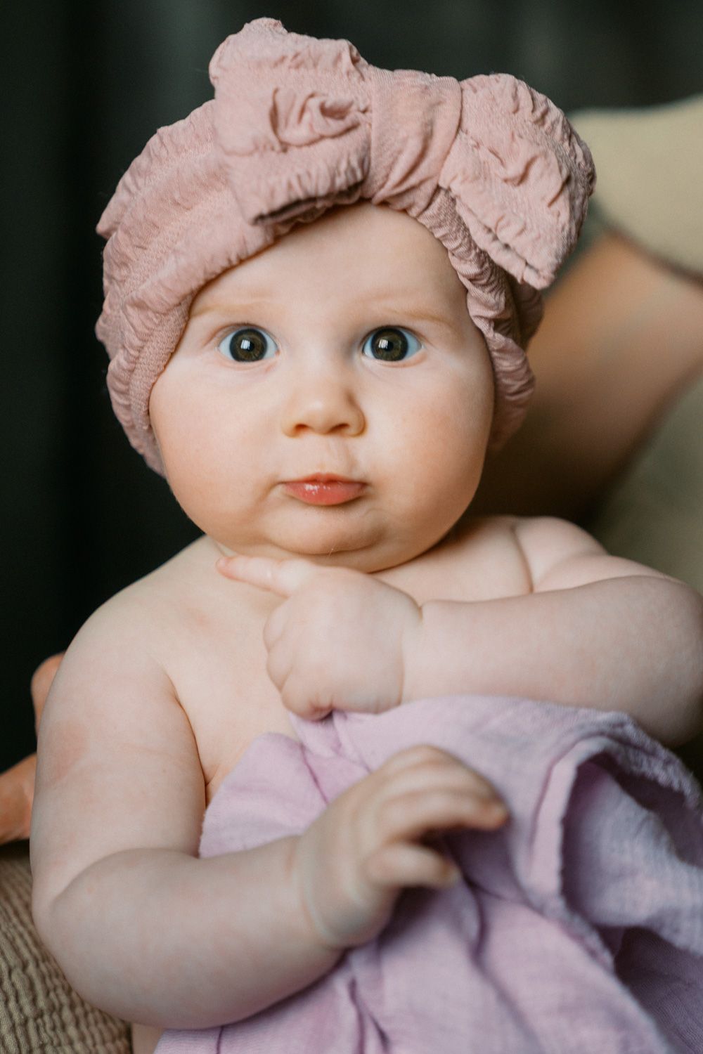Adorable baby girl wearing a pink bow headband, looking curiously at the camera while wrapped in a soft lavender blanket.