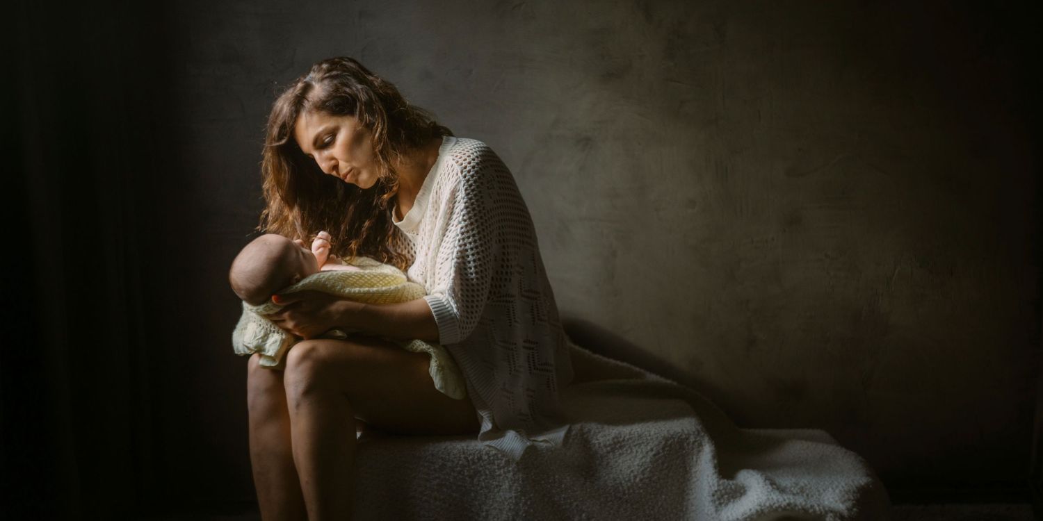 A mother tenderly holds her newborn baby in soft, moody lighting against a dark background.