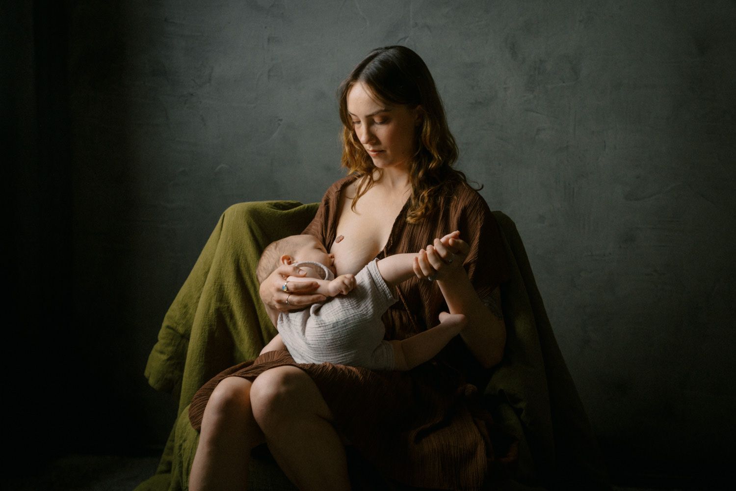 A mother tenderly breastfeeds her newborn baby while seated, draped in earthy brown and green fabric.