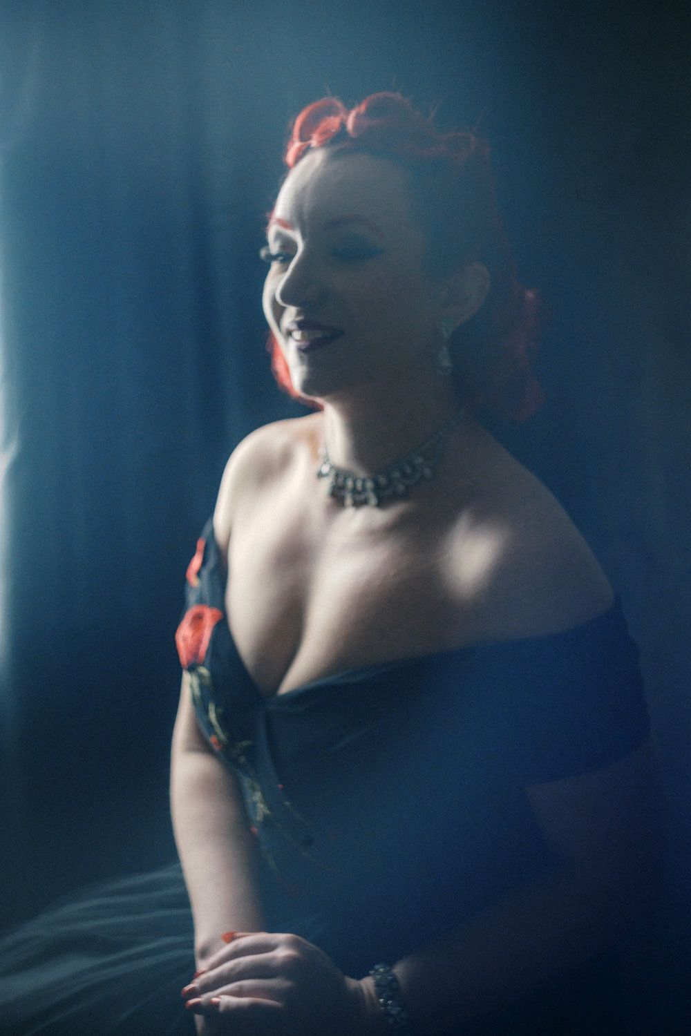 Woman in off-shoulder navy gown with red hair accessories and diamond necklace smiling in dramatic blue-lit portrait.