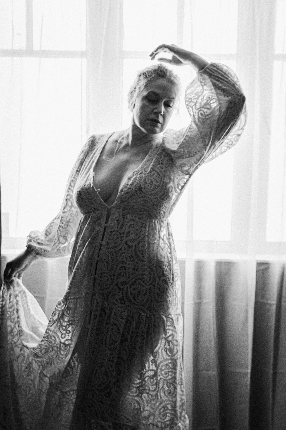 Black and white boudoir photo of woman in lace robe posing gracefully by bright window.