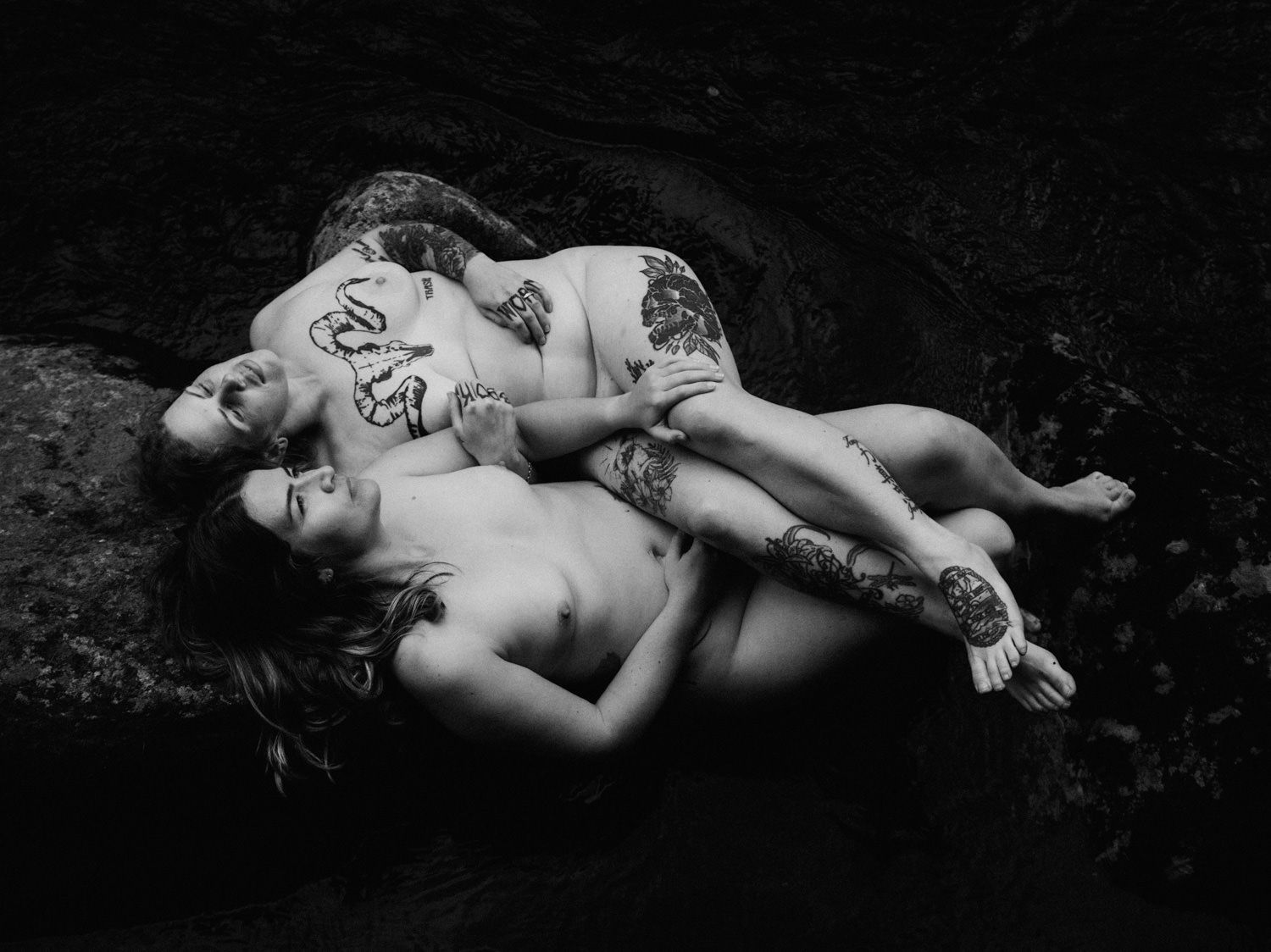 Black and white photo of two tattooed people resting on rocks in an artistic fine art portrait.
