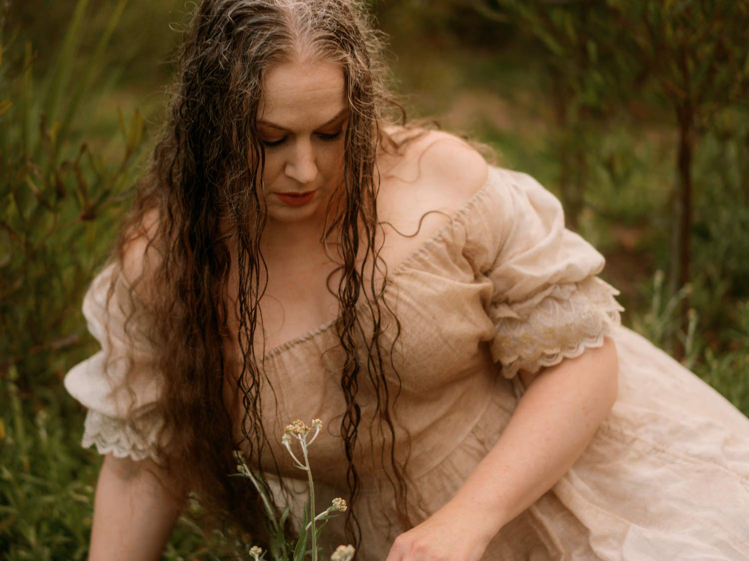Woman in vintage lace dress with wet curly hair picking wildflowers in a lush green meadow.