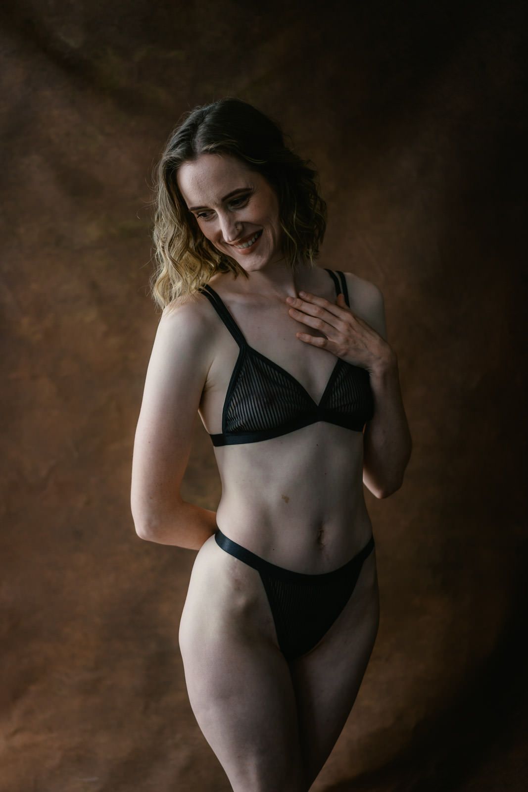 Woman smiling in black mesh lingerie set, posing against a warm brown textured studio background.