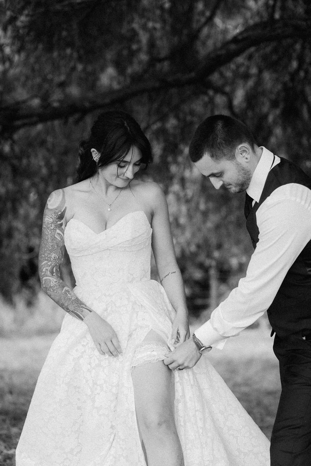 Black and white wedding photo of bride in lace gown with tattoos and groom adjusting her dress outdoors.