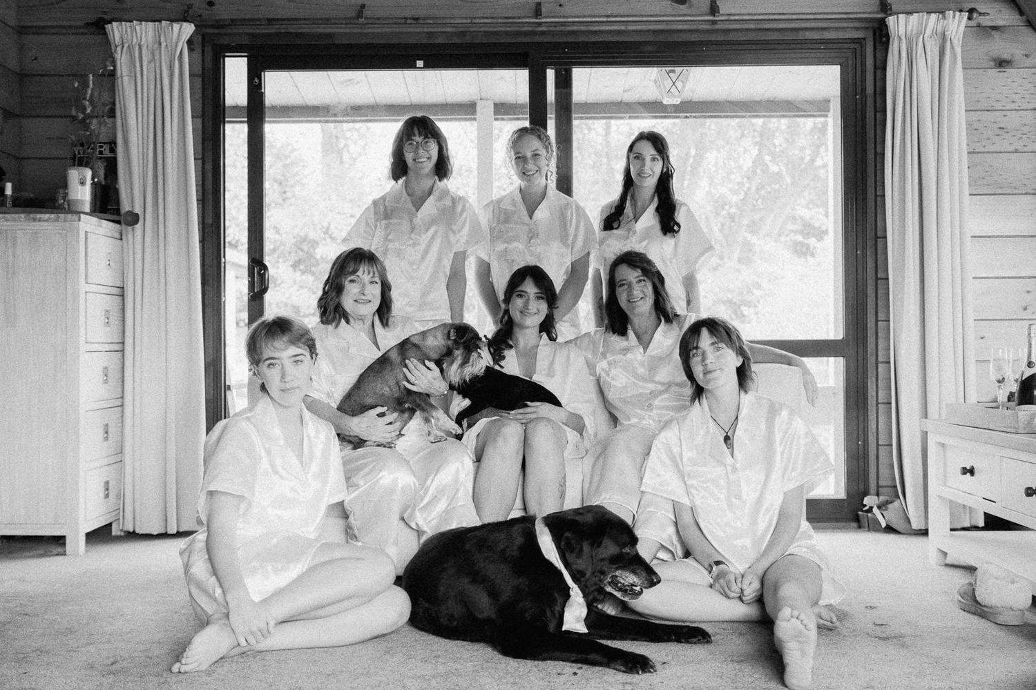 Black and white photo of eight women in white shirts posing together with two dogs in a cozy cabin room.