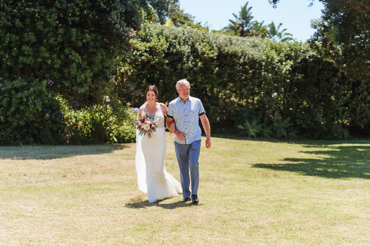 Bride in white gown walks with escort across green lawn toward outdoor wedding ceremony on sunny day.