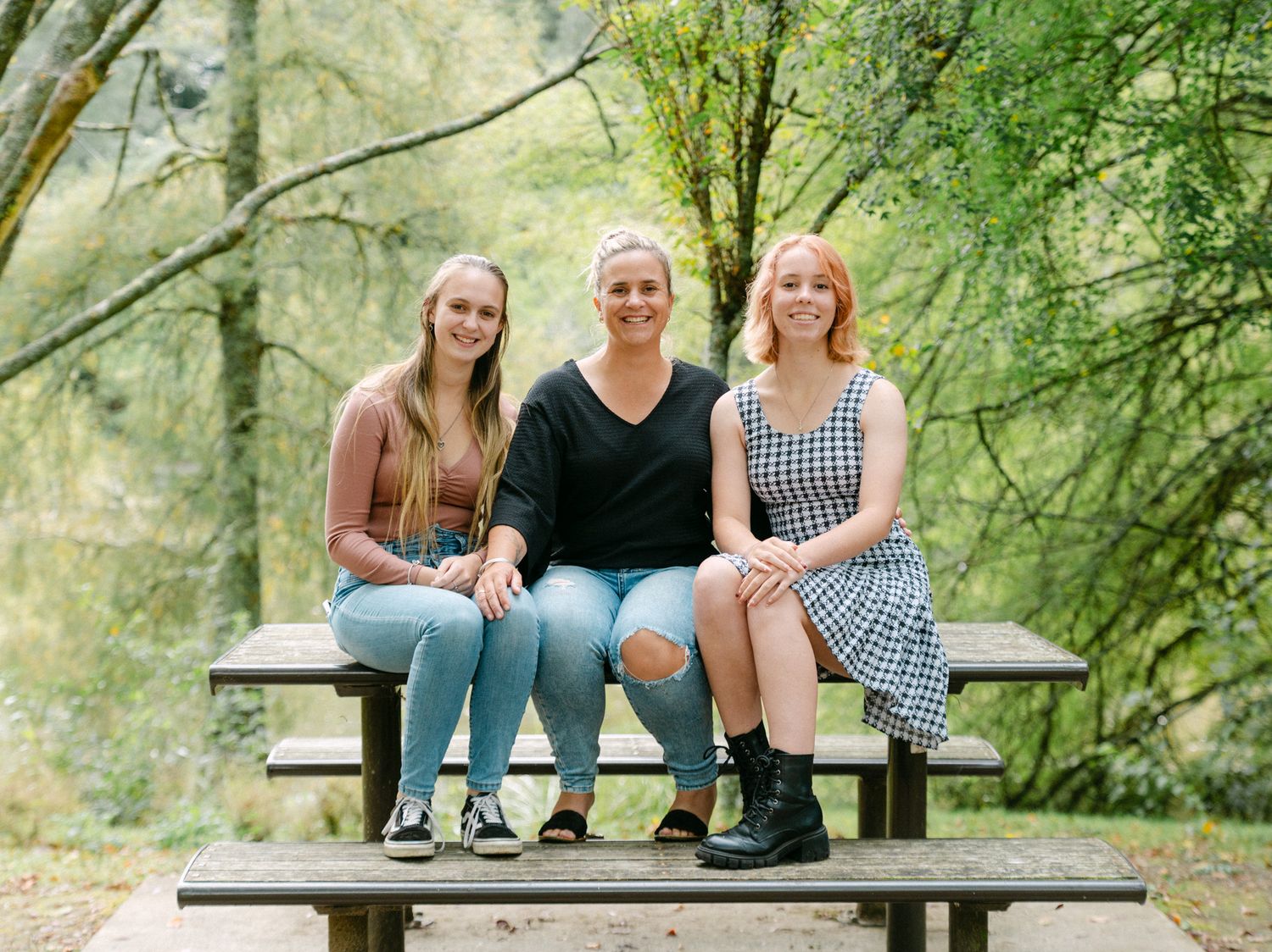 Three women smiling and sitting together on a picnic table bench outdoors in a lush green forest setting.