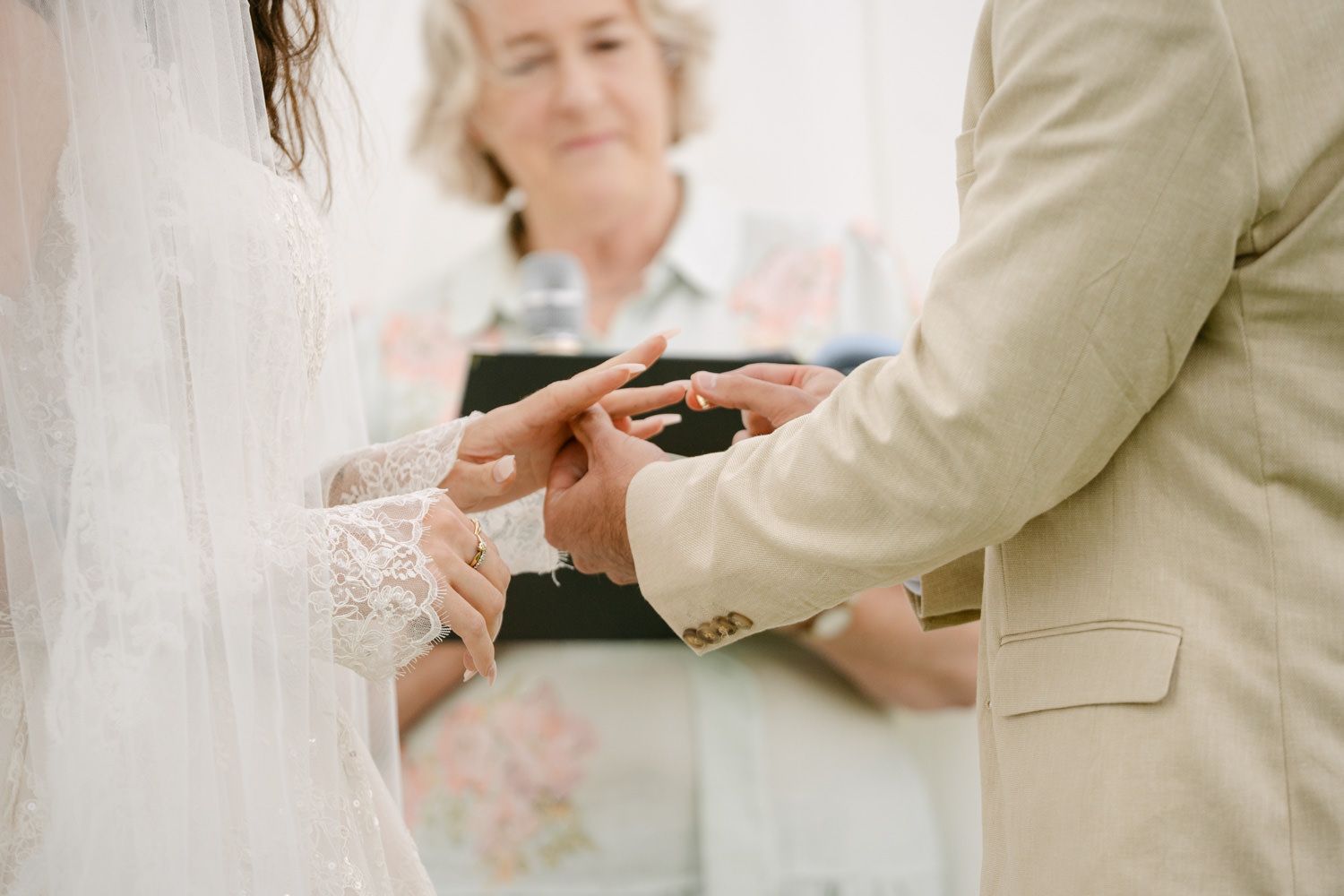 Groom placing wedding ring on brides finger during ceremony, officiant holding book in background.