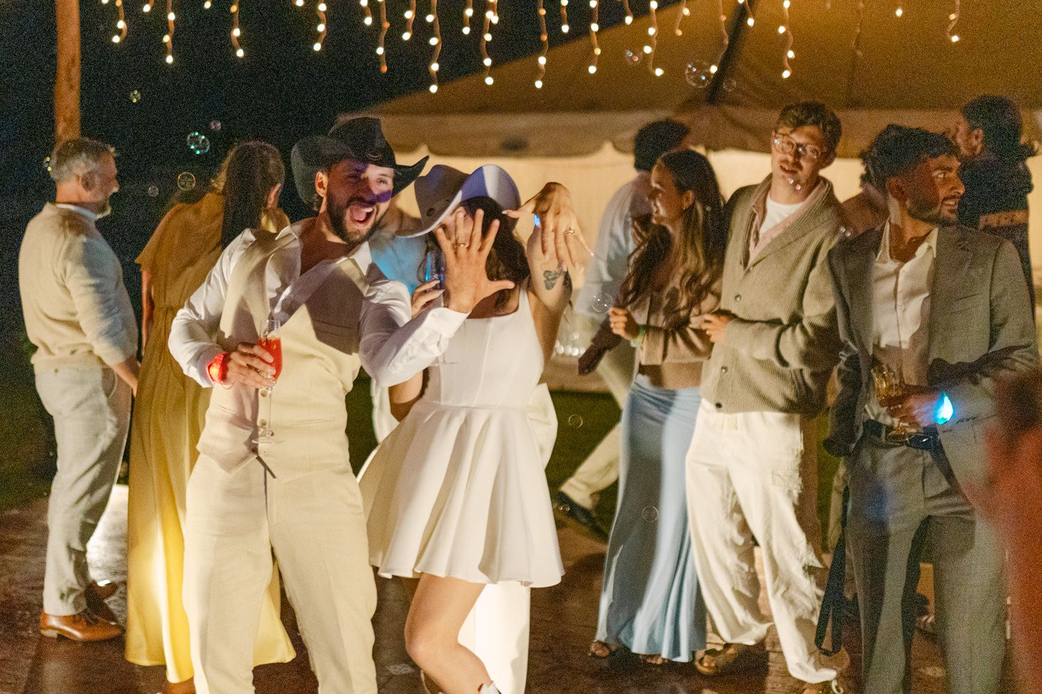 Couples dancing joyfully at an outdoor evening wedding reception under string lights in white attire.