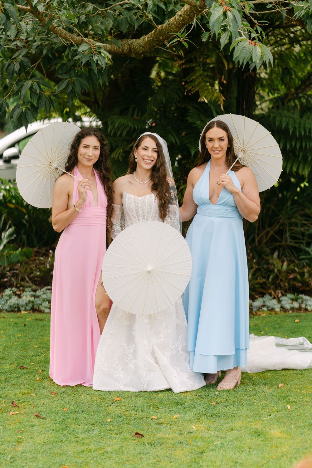 Bride in white gown with two bridesmaids in pink and blue dresses holding white parasols outdoors.
