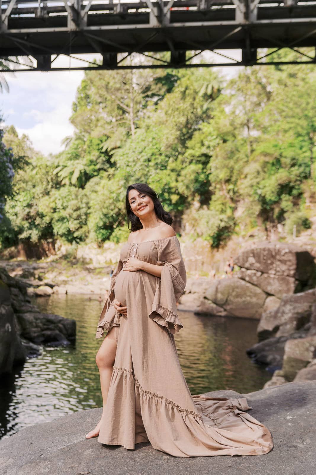 Woman in elegant beige ruffle gown posing by a serene river with lush greenery and a bridge backdrop.