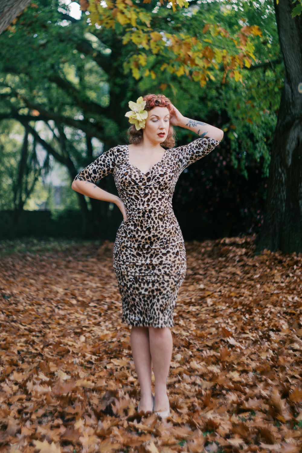 Woman in leopard print dress with floral hair accessory standing among autumn leaves in a lush green park.