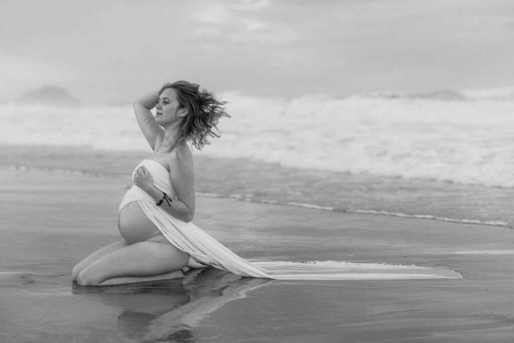 Black and white photo of a pregnant woman kneeling on a beach by the ocean.